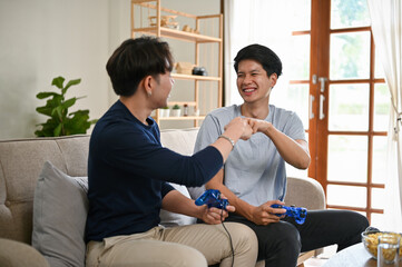 Two cheerful Asian male friends giving fist bump to each other while playing video games