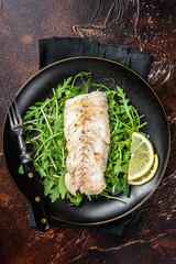 Grilled cod fish fillet served with green salad in a plate. Dark background. Top view