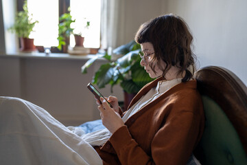 Pensive girl with eyeglasses lying resting on bed after college, using phone, chatting with friends, scrolling through social network, reading online book, watching movies. Procrastination concept