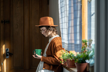 Pensive girl stands by window sadly look down thinking about relationships holding cup tea in hand. Tired young woman creative freelancer take coffee break from work after completing difficult project