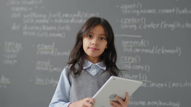 Slowmo portrait of elementary school Hisanic girl standing in front of blackboard in classroom holding tablet during programming lesson at daytime