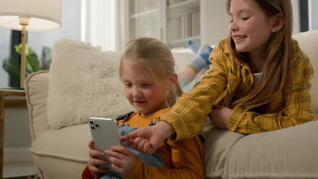 Two funny happy caucasian kids girls sisters friends children having fun playing on phone in living room couch play mobile online game together using smartphone browsing social network in Internet