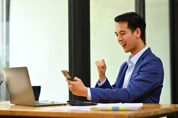 Surprised businessman looking at smart phone excited by good news online, celebrating success raising hands up and screaming