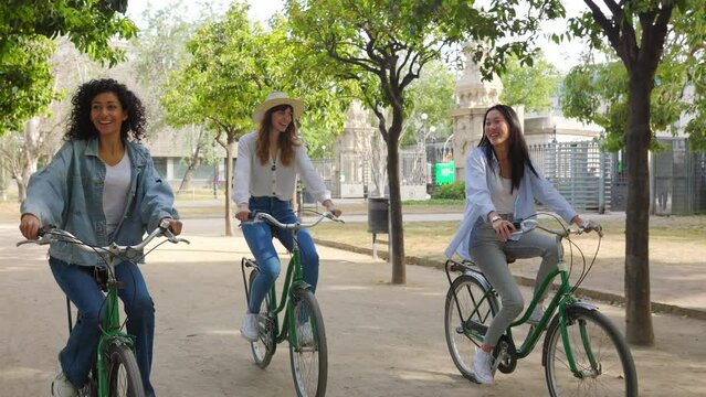 Young group of three multiracial women enjoying bicycle ride in city park. Diverse female friends having fun together riding bike during summer vacation outside. Youth community and travel concept.