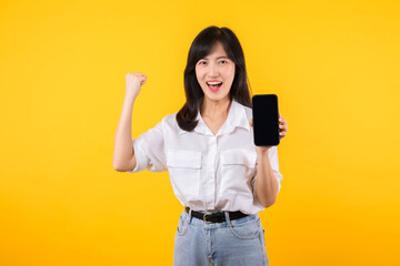 Portrait beautiful asian businesswoman happy woman wearing white shirt and denim plants showing blank smartphone screen and winner success body gesture feeling with fist up against yellow background.