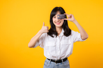 Asian young woman holding credit card in front of one eye and showing thumb up hand gesture with...