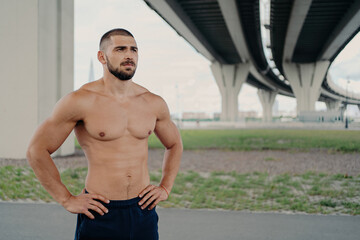 Obraz premium Shirtless bearded man poses outdoors, hands on waist, focused on distance, proud of perfect abs, sport enthusiast.
