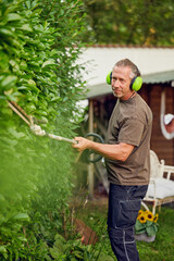 Gardener trimming a hedgerow using a hedge trimmer in the garden of a customer with earmuffs on for protection