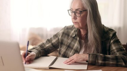 Focused gray haired senior woman studying online at home Curious mature female make notes in notebook while looking at laptop computer screen Distance remote education for person on pension