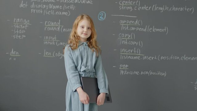 Slowmo medium portrait of Caucasian elementary age girl coming to blackboard holding laptop in hands in programming classroom at daytime