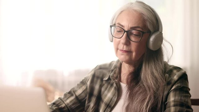 Portrait of senior woman with headphones listening to music while relaxing at home or working online browsing products in internet store reading news checking email on laptop computer indoors