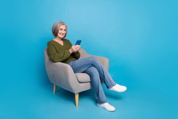 Fototapeta na wymiar Full size photo of positive cheerful person wear knit pullover sit on armchair typing sms on smartphone isolated on blue color background