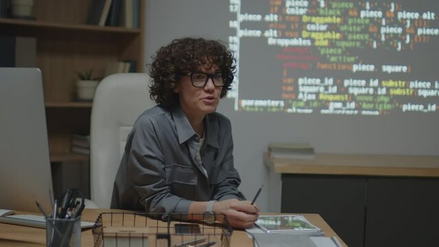 Medium shot of cheerful Caucasian female teacher sitting at desk in classroom pointing at screen projection with code explaining it