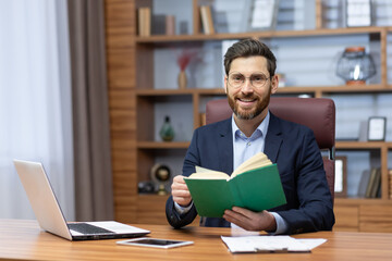 Joyful businessman in home office resting on break between work, mature man reading book, portrait of smiling boss holding book smiling and looking at camera.