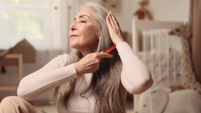 Portrait of beautiful senior woman looking at mirror and combing her long gray hair with wooden brush in the morning at home Concept of self care and daily routine