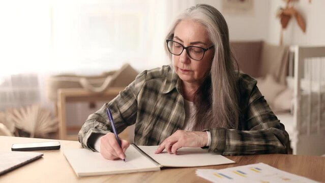 Focused gray haired senior woman with lack of new ideas or creative block writing and crossed out at notebook while finding solution idea correct answer for her question at cozy home