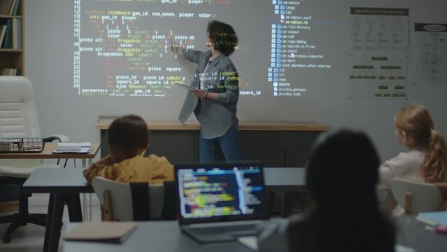 Full shot of Caucasian female teacher standing in front of screen projection pointing at it and explaining code to elementary age students
