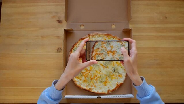 Blogger photographing fresh 4 cheese pizza. Top view - Female hands in blue sweatshirt use smartphone, take photo for social networks appetizing fresh cooked italian Quattro Formaggi Pizza in delivery