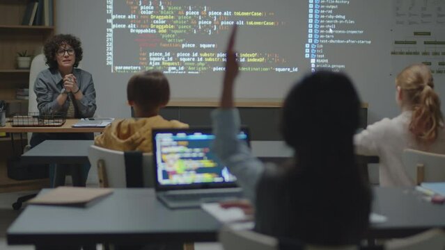 Over-the-shoulder full shot of Caucasian female teacher sitting at desk in classroom asking student raising hand to answer during programming lesson
