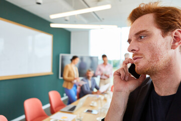Redhead businessman talking on mobile phone while sitting in front of colleagues discussing at office