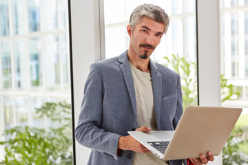 Fototapeta na wymiar Portrait of confident businessman holding laptop standing against window at office