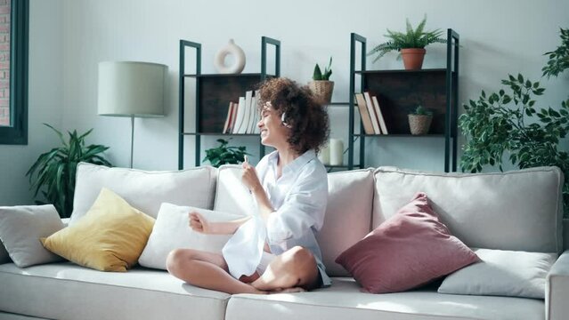 Video of motivated funny woman listening to music with headphones while dancing on couch at home