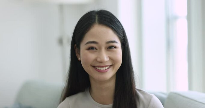 Head shot close up of happy beautiful single Asian young woman smile look at camera seated on couch at home, video call participant profile picture, relaxation at own or rented flat. Tenant portrait