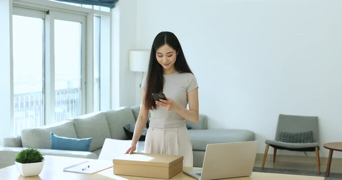 Young Asian woman prepares parcel box for sending to client hold cellphone ordering delivery courier services using website or mobile application. Shipment, business, modern tech, easy goods return