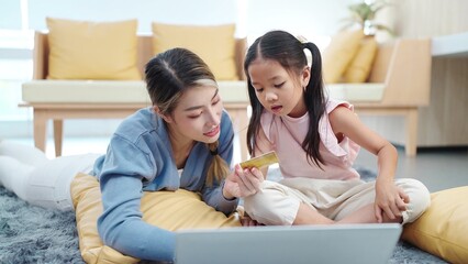 Happy asian mother and little daughter holding credit card for making online shopping on laptop while lying on floor in living room enjoying leisure together. Online shopping concept