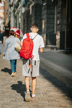 Young studen guy with backpack walking in city, holding smartphone and listening music headphones, rear view