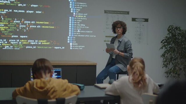Full shot of female Caucasian teacher sitting on chair in front of elementary age students showing them code on screen projection and giving explanations