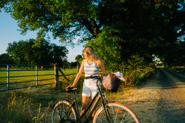 Young attractive caucasian woman with bicycle near horses pasture in summer day at sunset.