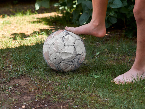 Horizontal photo. Close-up of a young girl's barefoot soccer player's bare feet on an old and battered soccer ball.