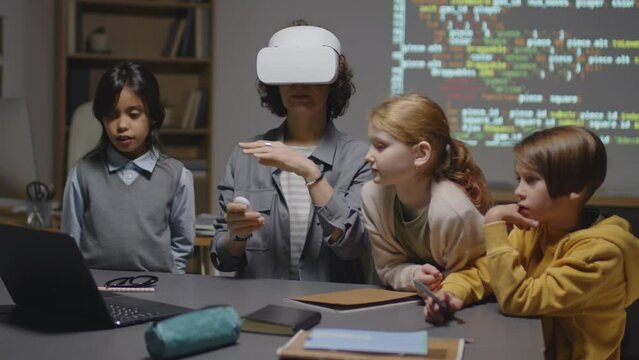 Medium full shot of female Caucasian teacher in VR goggles sitting at desk working with group of ethnically diverse students listening to her