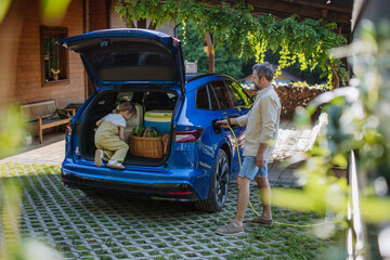 Father charging their electric car, little daughter sitting and waiting in a car trunk.