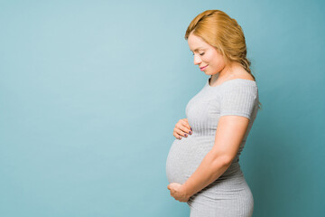 Cute pregnant woman in her 30s looking at her belly and looking happy in a studio with blue...
