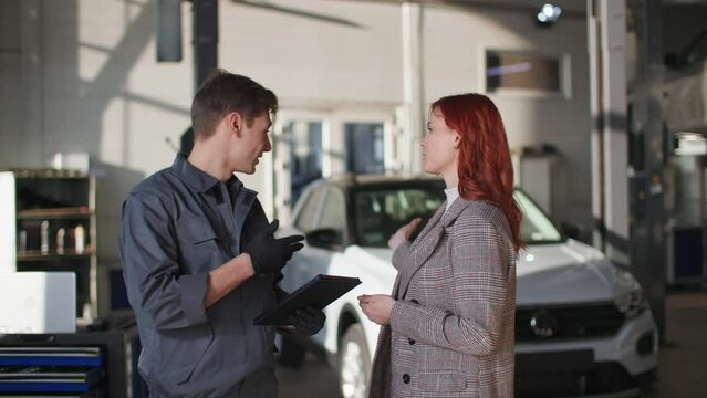 automobile maintenance, male mechanic uses modern technology and shows technical moment of car on tablet beautiful female client, smiles and looks at camera