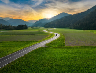 Top drone view of beautiful landscape with alpine mountains, paved highway, hills, fields green grass. Aerial view of beautiful curved rural road in green meadows at sunset in summer in Slovenia