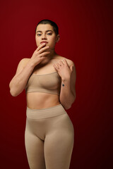Fototapeta premium body love, young tattooed woman in beige underwear posing on red background, body positivity, curvy fashion, comfortable in skin, curvy model, generation z, self love, looking at camera