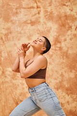 denim fashion, gen z, happy curvy woman with tattoo posing on mottled beige background, body positivity movement, self-esteem, confidence, short haired model, youth culture