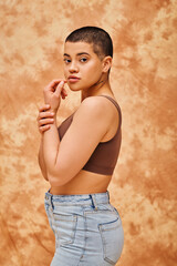 Obraz premium representation of body, curvy and young woman in crop top and jeans posing on mottled beige background, short haired, self-acceptance, generation z, tattooed, different shapes, looking at camera