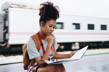 Asian teenage girl african american traveler dressed in casual wear holding map and searching right direction of route setting while waiting for a train at the station.