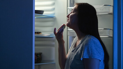 Woman opens refrigerator and looks at tempting donuts with chocolate cream at night. Brunette lady wants to eat tasty donuts during midnight - obrazy, fototapety, plakaty