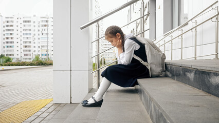 Fototapeta na wymiar Brown-haired girl sits on empty entrance staircase without desire to attend lessons at school. Junior schoolgirl sits with sad expression