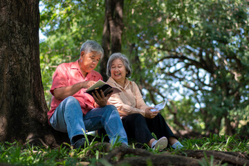 Happy Asian old senior couple with gray hair reading a book outside in park. Concept of happy elderly after retirement and good health