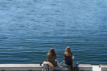 Zwei junge Frauen mit langen Haaren sitzen auf einer langen Bank am Wasser und essen und trinken in...