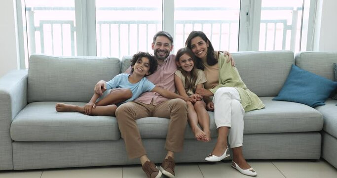 Loving attractive multinational family sit on couch in living room look at camera and posing, smile, feel happy, have home funny weekend together, parents demonstrate love, care and warm relationship
