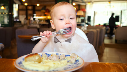 Toddler girl likes to eat food by licking large fork in cozy restaurant. Daughter enjoys eating meal independently in crowded cafe at resort