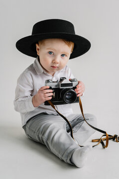 Fun kid holds retro vintage camera isolated on white wall. Child in a hat sitting on floor and play with photo camera. International Photographers Day. Children's studio portrait. Mockup. Close Up.