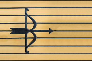 Cast iron in the form of a bow with an arrow fixed on a metal grate on a plain ocher background.